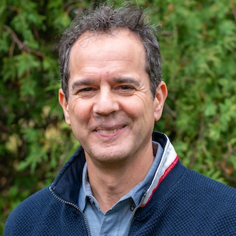 The image shows a middle-aged man with short, graying hair and a friendly expression. He is wearing a blue jacket over a blue shirt. The background is a blurred green, suggesting an outdoor setting, possibly a garden or park. The man appears to be smiling, and the overall impression is one of approachability and warmth.